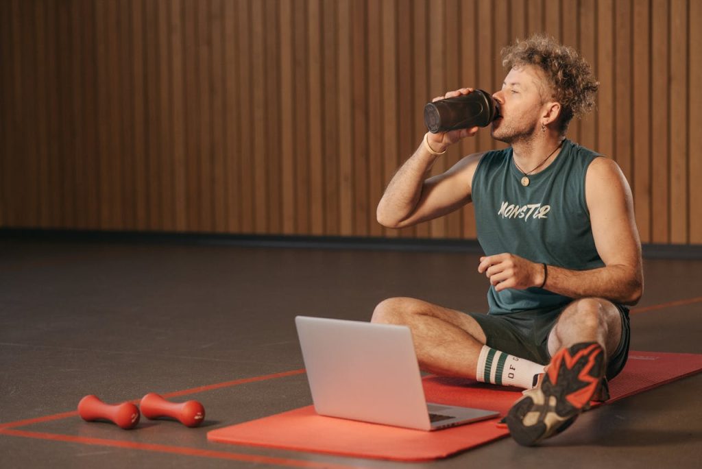 man-drinking-from-a-tumbler-6339725 Man in gym drinking water during workout on exercise mat with laptop.