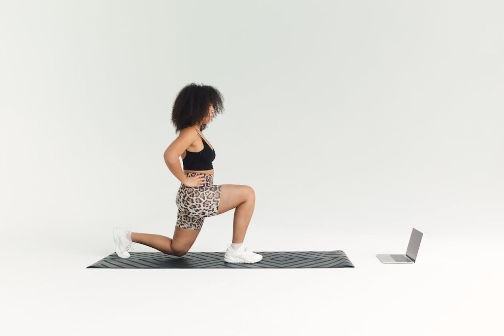 woman-watching-online-workout-video-6516232 A woman doing a lunge exercise indoors with a laptop for guidance.